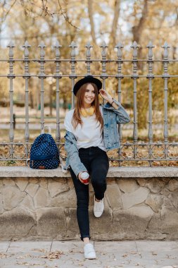 Parkta şapka, sırt çantası ve sarı kulaklık takan kahverengi saçlı güzel, gülümseyen hipster kız öğrenciportresi.