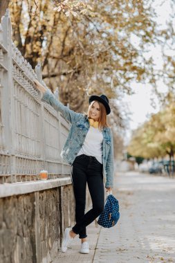 Parkta şapka, sırt çantası ve sarı kulaklık takan kahverengi saçlı büyüleyici, gülümseyen hipster kız öğrenciportresi.