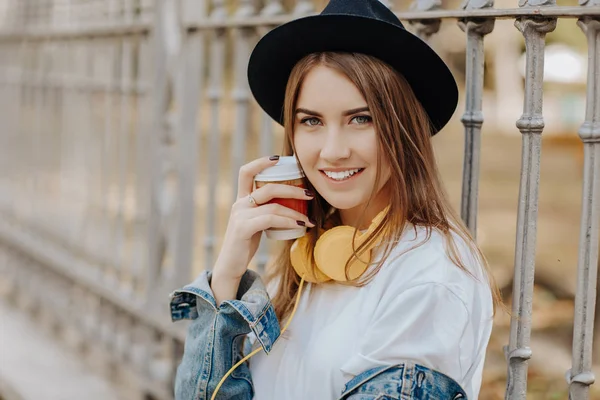 Photo of a beautiful smiling hipster girl with brown hair wearing a hat and holding cup of tea or coffee in the park — Stock Photo, Image