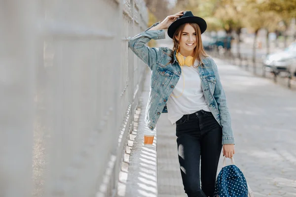 Retrato de una hermosa estudiante hipster sonriente con el pelo castaño usando un sombrero, mochila y auriculares amarillos en el parque . — Foto de Stock