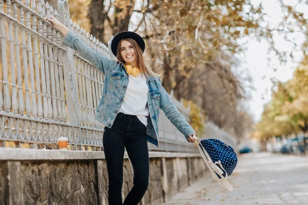 Parkta şapka, sırt çantası ve sarı kulaklık takan kahverengi saçlı büyüleyici, gülümseyen hipster kız öğrenciportresi.