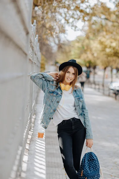 Retrato de uma bela estudante hipster sorridente com cabelo castanho usando um chapéu, mochila e fones de ouvido amarelos no parque . — Fotografia de Stock