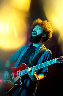 Neon ışıklar kırmızı gitar ile kıvırcık saçlı genç hipster adam. Rock müzisyeni elektrik gitar çalıyor.