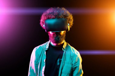 Neon ışıklar stüdyoda sanal gerçeklik gözlüğü giyen kıvırcık saçlı genç sakallı hipster adam Fotoğraf. Vr kulaklıkla akıllı telefon kullanma.