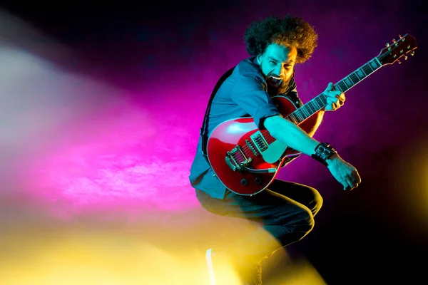 Jonge hipster man met krullend haar springen met rode gitaar in neon lichten. Rock muzikant speelt elektrische gitaar. — Stockfoto
