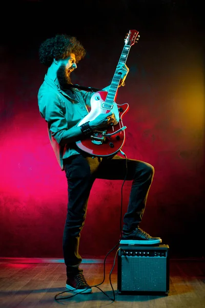 Portrait of hipster man with curly hair with red guitar in neon lights. Rock musician is playing electrical guitar. — Stock Photo, Image