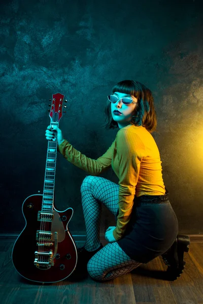 Beautiful young hipster woman with curly hair with red guitar in neon lights. Rock musician is playing electrical guitar. 90s style concept. — Stock Photo, Image