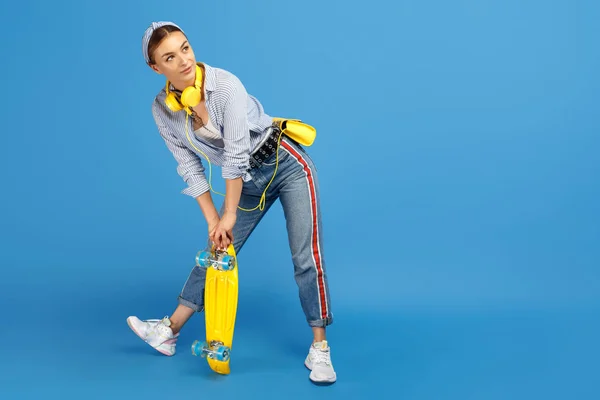 Полная фотография довольно беззаботной молодой женщины с жёлтыми наушниками, позирующей с пенни или скейтбордом на синем фоне . — стоковое фото