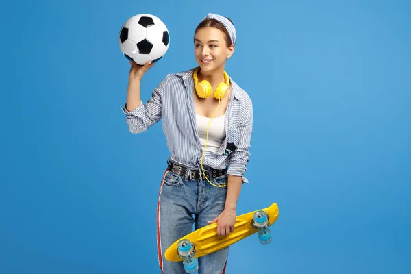 黄色のペニーやスケートボード、サッカーボール、青い背景の上にポーズをとるヘッドフォンを持つ愛らしい若い女性の写真. — ストック写真