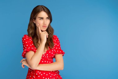 Kırmızı elbiseli odaklanmış genç kadın mavi arka plan üzerinde ciddi bir yüz ile düşünmek uzağa bakıyor.