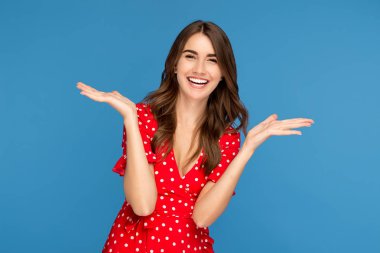 Mutlu genç bir kadın, parlak bir gülümseme içinde, kırmızı bir elbise içinde heyecanlı bir yüzle, mavi arka planda elleri havada yukarı bakıyor..