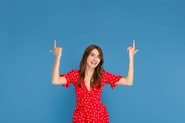 Mavi arka plan üzerinde parmaklarını işaret kırmızı elbise parlak gülümseme ile Çekici genç kadın.