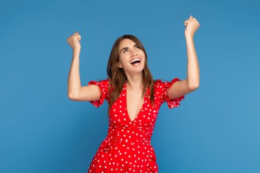 Mutlu genç bir kadın, parlak bir gülümseme içinde, kırmızı bir elbise içinde heyecanlı bir yüzle, mavi arka planda elleri havada yukarı bakıyor..
