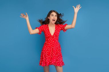 Mavi arka plan üzerinde elleri ile heyecanlı yüzü ile kameraya bakarak kırmızı rahat elbise parlak gülümseme ile Muhteşem genç kadın.