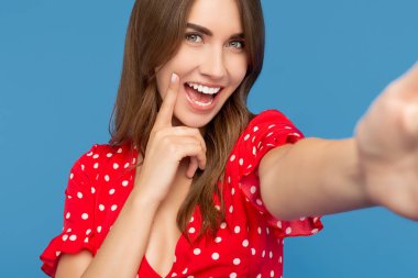 Mavi arka plan üzerinde onu beyaz dişleri gösteren kamera bakarak kırmızı rahat elbise parlak gülümseme ile Neşeli genç kadın. Kadın akıllı telefon ile selfie çekmek.