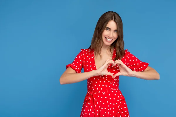 Mujer joven bonita con sonrisa brillante en vestido rojo muestran gesto de amor sobre fondo azul. Concepto del Día de San Valentín . — Foto de Stock