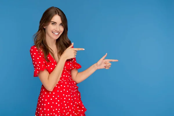 Mujer joven divertida con sonrisa brillante en vestido rojo señalando los dedos sobre fondo azul . —  Fotos de Stock