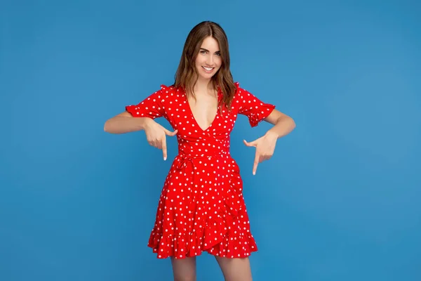 Mulher muito jovem com sorriso brilhante em vestido vermelho apontando os dedos para baixo sobre fundo azul . — Fotografia de Stock