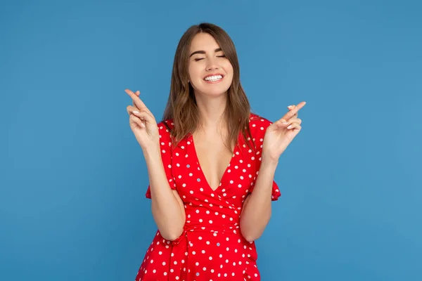 Boldog nő, világos mosoly a piros ruhában vezetése szemek zárt kezével ujjak keresztbe a remény a kék háttér. — Stock Fotó
