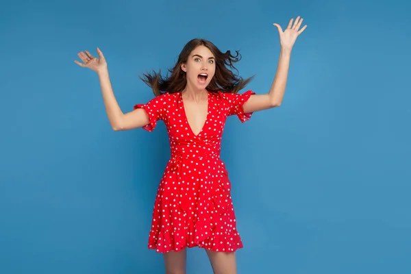 Linda jovem com sorriso brilhante em vestido casual vermelho olhando para a câmera com o rosto animado com as mãos para cima sobre o fundo azul . — Fotografia de Stock