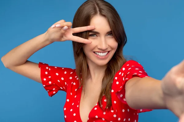 Mujer joven feliz con sonrisa brillante en vestido rojo tomar selfie con teléfono inteligente que muestra el gesto de paz sobre fondo azul . —  Fotos de Stock