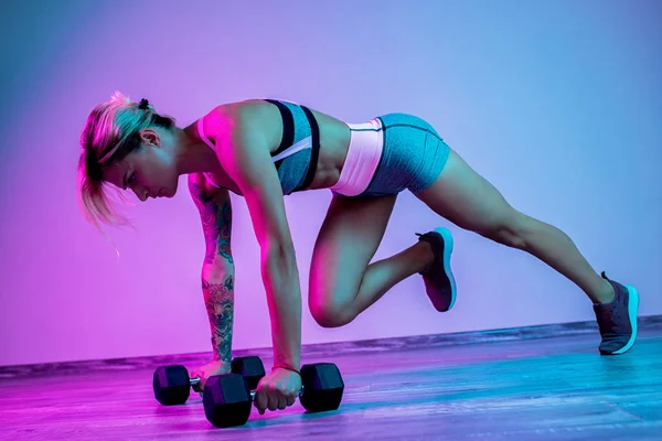 Atletische jonge blonde vrouw doet zware halter oefening in de sportschool in neonlichten. — Stockfoto
