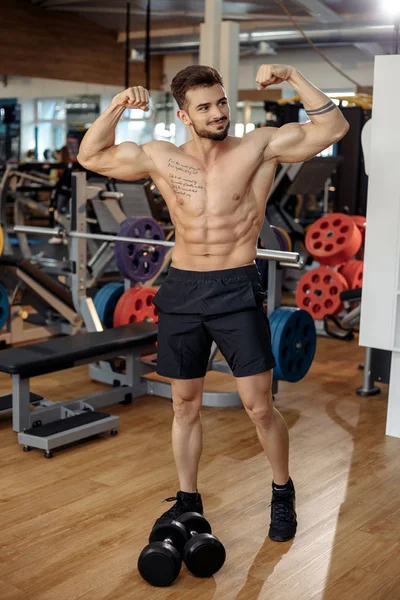 Izmos sportos fiatalember testépítő fitness modell megmutatja a bicepsz után él a súlyzó tornaterem. — Stock Fotó