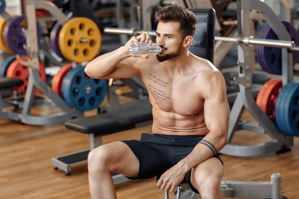 运动健美的年轻人健美模特儿在健身房的长椅上用杠铃锻炼后喝水. — 图库照片