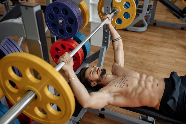 强壮的运动青年男子健美健身模型锻炼胸部与杠铃在健身房的长凳上。顶部视图. — 图库照片