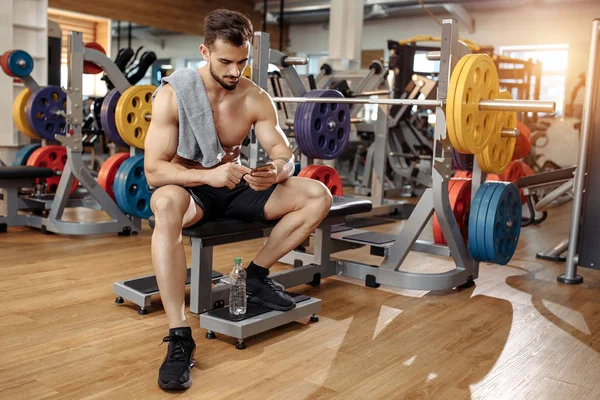 Atractivo atlético joven culturista modelo de fitness utilizar smartphone o teléfono móvil en el banco en el gimnasio. —  Fotos de Stock