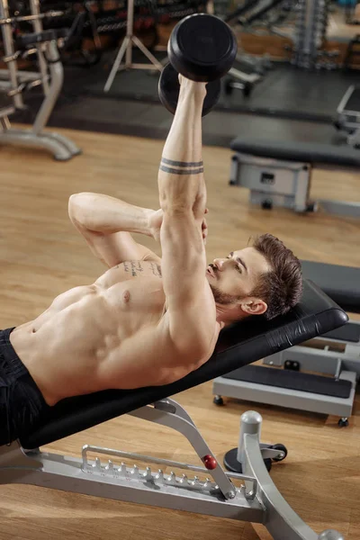 Muscular atlético jovem fisiculturista modelo de fitness exercício tríceps com halteres no banco de ginástica . — Fotografia de Stock