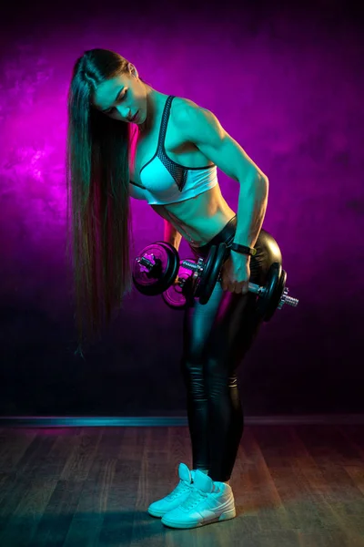 Motivated young woman fitness model workout with professional dumbbells in neon lights in the studio.
