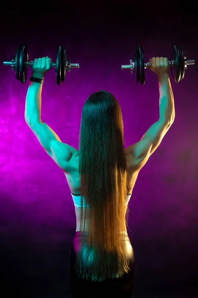 Motivated young woman fitness model exercise shoulders with professional dumbbells in neon lights in the studio. Back view.