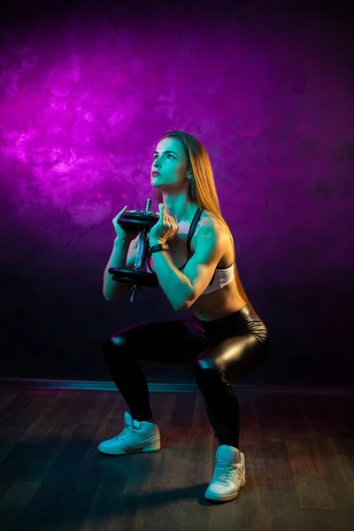 Focused young woman fitness model doing squats with professional dumbbells in neon lights silhouette in the studio. — Stock Photo, Image