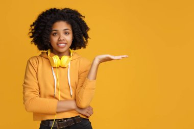Parlak gülümseme ile yakışıklı afrikalı-amerikalı genç kadın rahat giysiler ve kulaklık lar içinde giyinmiş copyspace ya da sarı arka plan üzerinde bir şey gösteren giyim.