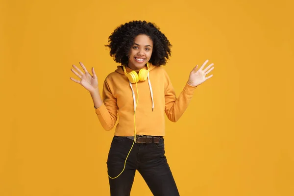 Schöne afrikanisch-amerikanische junge Frau mit strahlendem Lächeln in lässiger Kleidung und Kopfhörer bereit für Umarmungen vor gelbem Hintergrund. — Stockfoto