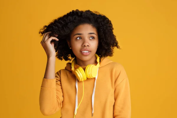 Charmante afrikanisch-amerikanische junge Frau in lässiger Kleidung und Kopfhörer, die über gelben Hintergrund nach oben schaut. — Stockfoto