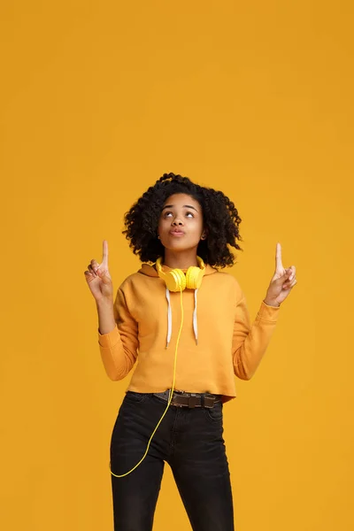 Charmante afrikanisch-amerikanische junge Frau mit strahlendem Lächeln in lässiger Kleidung und Kopfhörer-Kleidung, die Finger nach oben über gelbem Hintergrund zeigt. — Stockfoto