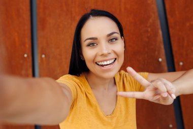 Akıllı telefon selfie yapıyor rahat giysiler giymiş parlak bir gülümseme ile Çekici esmer genç kadın ve paslı duvar önünde barış jesti göstermek.