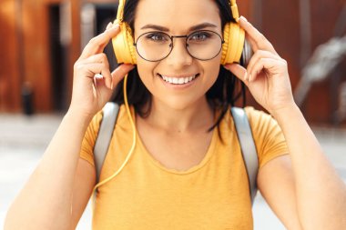 Sarı kulaklıkile müzik dinleyerek ve sokakta gülümseyen rahat giysiler giymiş parlak bir gülümseme ile Pretty esmer genç kadın öğrenci.