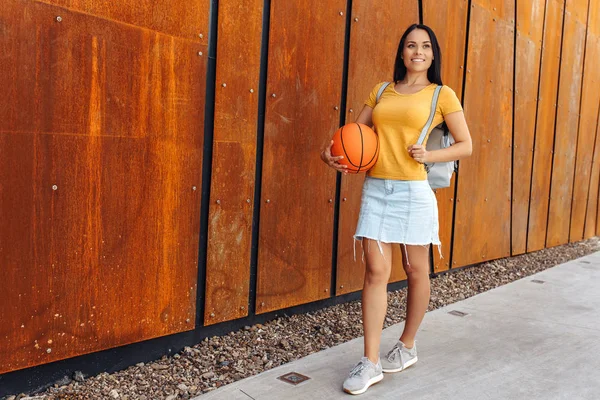 Mooie brunette jonge vrouw student met heldere glimlach gekleed in Casual moderne kleren poseren met mand bal en stijlvolle rugzak voor Rusty muur. — Stockfoto