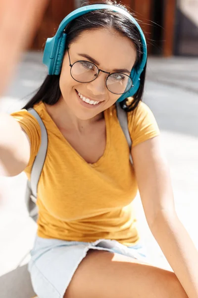 カジュアルな服に身を包んだ明るい笑顔の面白いブルネットの若い女性は、通りでスマートフォンで自分撮りを取ります. — ストック写真