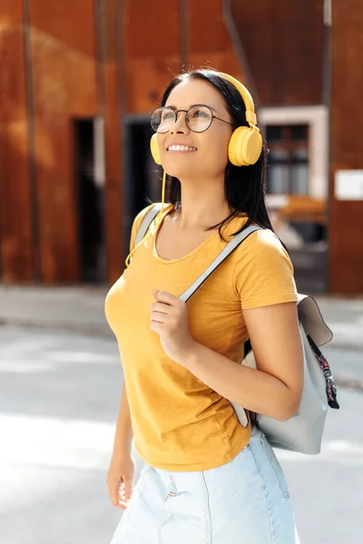 黄色のヘッドフォンで音楽を聴き、通りで笑顔を見せるカジュアルな服を着た明るい笑顔の魅力的なブルネットの若い女子学生. — ストック写真