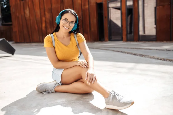 Brunette young woman student with bright smile dressed in casual clothes listening music on headphones while sitting on cement floor in front of rusty wall. — Stock Photo, Image