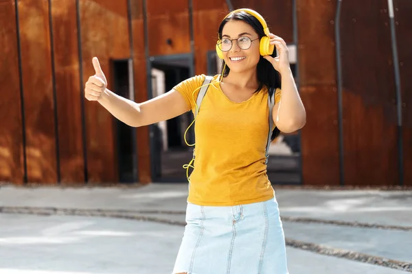 黄色のヘッドフォンで音楽を聴き、通りでOKジェスチャーを示すカジュアルな服を着た明るい笑顔のブルネットの若い女子学生. — ストック写真