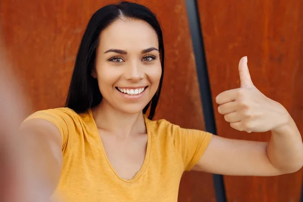 Jeune femme brune drôle avec un sourire lumineux habillé de vêtements décontractés faire selfie sur smartphone et montrer ok geste devant un mur rouillé . — Photo