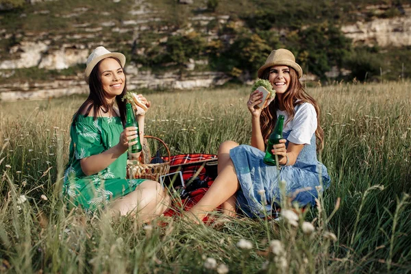 İki çekici genç kadın oturup piknik battaniye var aşağı ve eğlenin. Bira ve sandviç. — Stok fotoğraf