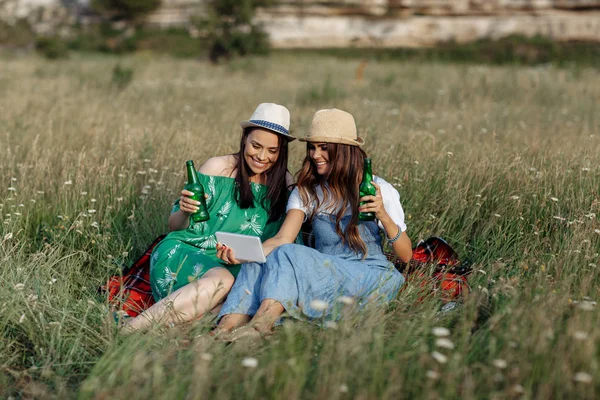 Deux jolies jeunes femmes assises sur la couverture de pique-nique et prendre selfie sur smartphone ou tablette. Bière et sandwichs . — Photo
