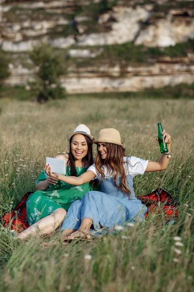 Deux jolies jeunes femmes assises sur la couverture de pique-nique et prendre selfie sur smartphone ou tablette. Bière et sandwichs . — Photo