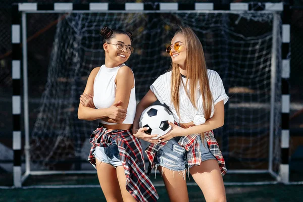 Δύο σύγχρονα νεαρά κορίτσια hipster ποζάρουν με χαρούμενα πρόσωπα μπροστά από το τέρμα στο γήπεδο ποδοσφαίρου. — Φωτογραφία Αρχείου